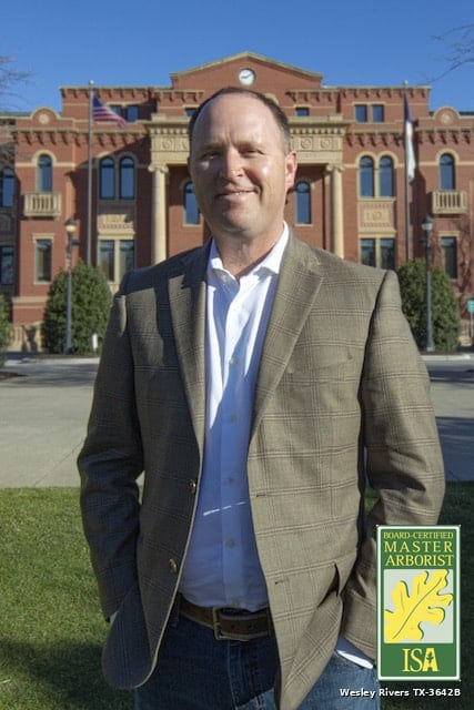 Wes Rivers, ISA Board-Certified Master Arborist, standing in front of a historic red brick courthouse, wearing a tan blazer over a white shirt.